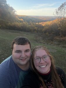 Two people standing outside, posing in front of a vista of autumn trees.