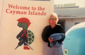 Kathy standing in front of a sign that says Welcome to the Cayman Islands.