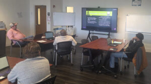 People sitting at tables in a training room looking at a large, flat panel monitor.