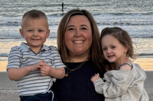 Emilee Sames standing on an ocean beach holding her daughter and son.