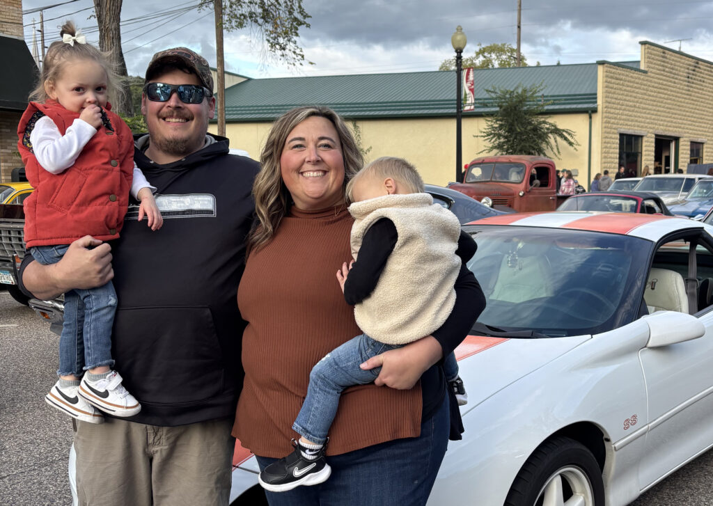 Emilee Sames with her husband holding their children and standing in front of a white car.