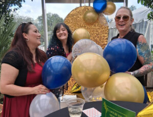 Three women standing and smiling while holding balloons during the celebration.