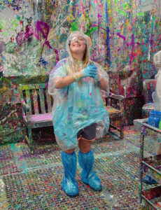 Young woman smiling and standing in the middle of a paint-splattered room wearing a clear plastic poncho and shoe coverings, all covered in multiple colors of paint.