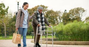 A woman caregiver walks with an older woman who is using a walker.