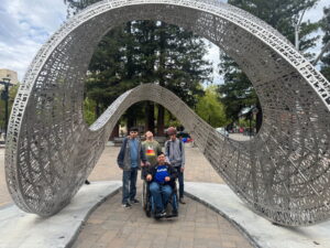 Four men outside in a park posing for picture underneath a large, oblong, metal sculpture.