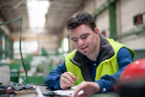 A young man sits at a table in a warehouse writing something on a clipboard.